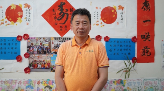 介護施設「一笑苑」内の壁に飾られた写真や書画の作品の前に立つ「一笑苑」代表取締役の佐々木弘志さん（撮影・許可）。