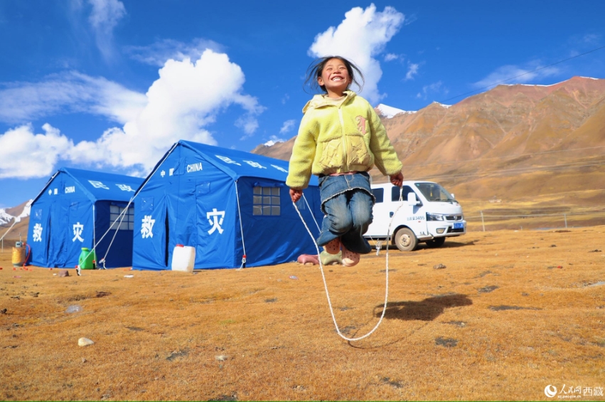 避難所のテントの前で、満面の笑みを浮かべながら縄跳びをする女の子。（撮影・次仁羅布）